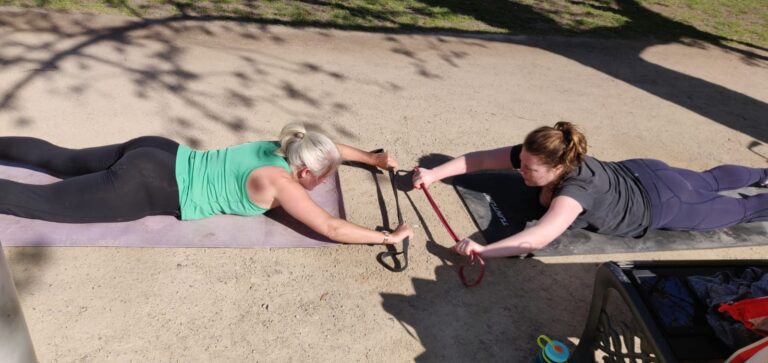 2 vrouwen in sportkleding trainen buiten in een park, doen een grondoefening met elastieken